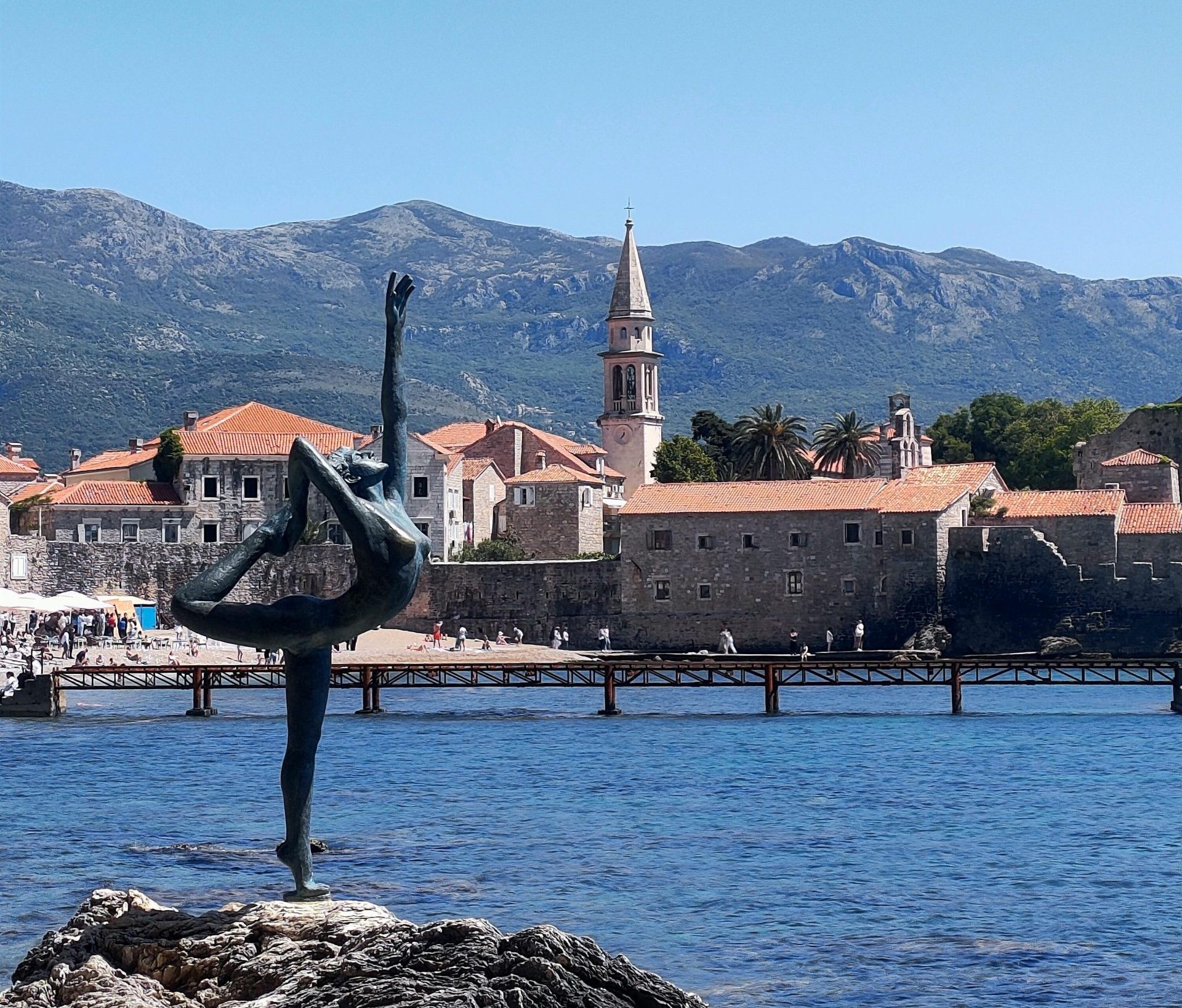 Sculpture of a dancer by the sea in Budva, Montenegro with a historic town and mountains in the background.