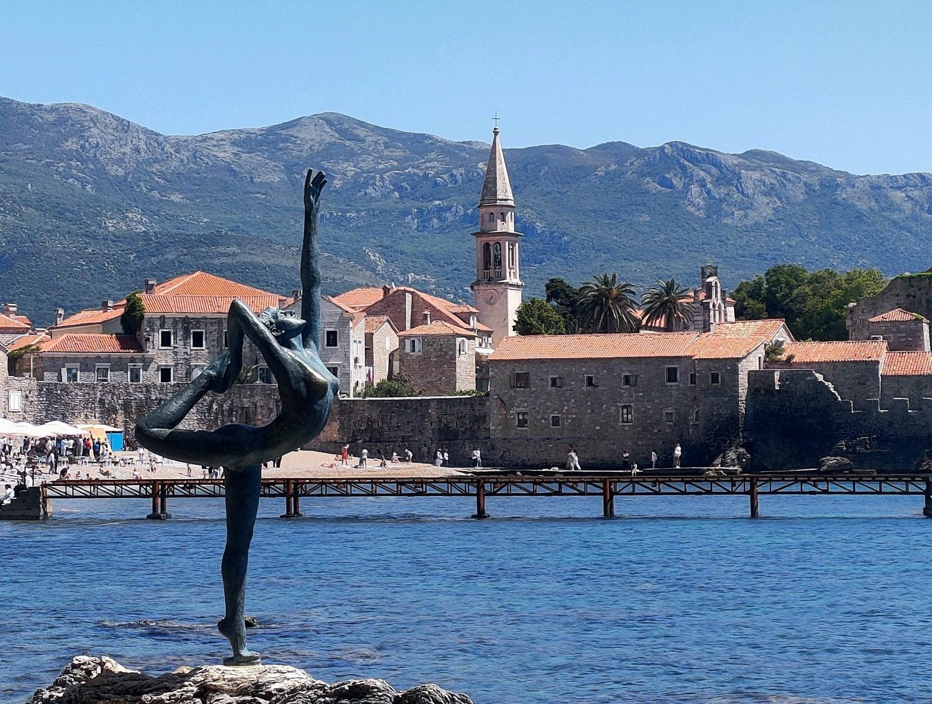 budvaoldtown Sculpture of a dancer by the sea in Budva, Montenegro with a historic town and mountains in the background.